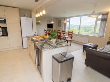A kitchen island with a plant bread and fruit bowl next to dining table and chairs in an open kitchen living room with large window showing a green landscape at The Gables in Sheepscombe