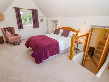 A bedroom with a double bed purple pillows and blanket an armchair near a window and a staircase at The Gables in Sheepscombe