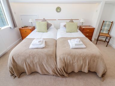 A bedroom with two single beds side by side each with pillows and folded towels on beige blankets between two wooden nightstands with lamps and a wooden chair with a plaid cushion at The Gables in Sheepscombe