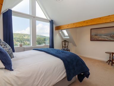 A bedroom with a bed dressed in white and blue near a large window with blue curtains and a wooden chair at The Gables in Sheepscombe