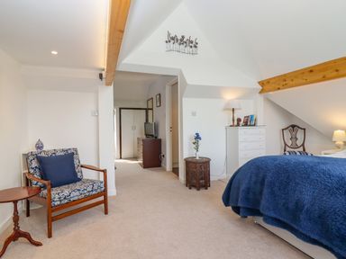A bedroom with a bed covered in a blue blanket a wooden chair with floral cushions a small round wooden table a white chest of drawers a decorative side table and a television at The Gables in Sheepscombe