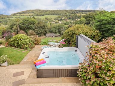 A garden with a hot tub, striped lounge chairs, and flowering bushes with hills in the background at The Gables in Sheepscombe