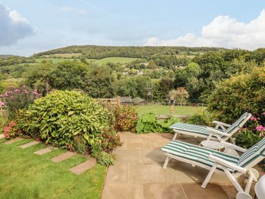 A garden patio with two striped lounge chairs surrounded by plants and a view of hills and trees at The Gables in Sheepscombe