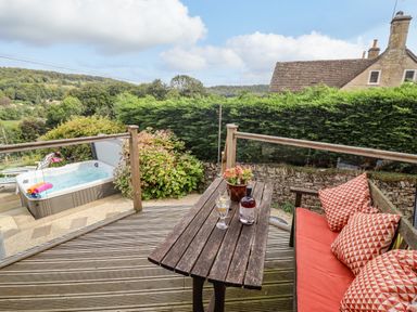 An outdoor wooden deck with a bench and table next to a hot tub and garden at The Gables in Sheepscombe