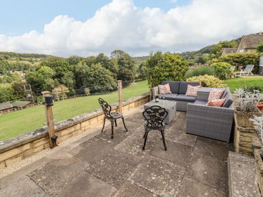 An outdoor patio with chairs and a sofa set overlooking a green field and hills at The Gables in Sheepscombe