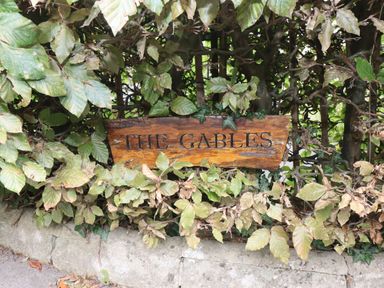 A wooden sign that says The Gables surrounded by green and brown leaves on a bush in Sheepscombe
