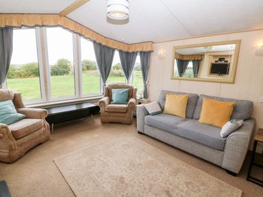 A living room with a grey sofa and two armchairs with cushions a large mirror on the wall and large windows with curtains at Plas Tirion Caravan Llanfairynghornwy near Cemaes Bay
