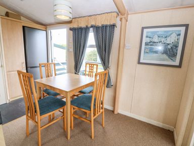 A dining area with a wooden table and four chairs near a window with curtains and a painting on the wall at Plas Tirion Caravan in Llanfairynghornwy near Cemaes Bay