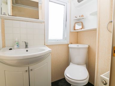 A bathroom with a white sink, a mirror, a window, a toilet, and shelves with small items at Plas Tirion Caravan in Llanfairynghornwy near Cemaes Bay