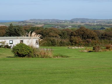 A green field with bushes and shrubs and a caravan with hills in the background at Plas Tirion Caravan Llanfairynghornwy near Cemaes Bay