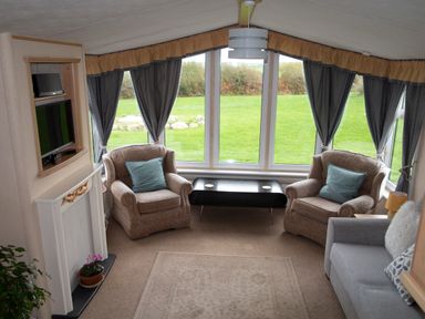 A sitting area with two armchairs and a sofa facing a low black table with large windows and curtains at Plas Tirion Caravan Llanfairynghornwy near Cemaes Bay