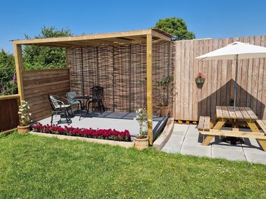 An outdoor seating area with a pergola, chairs, table, wooden picnic table with umbrella, and flower pots at Plas Tirion Caravan in Llanfairynghornwy near Cemaes Bay