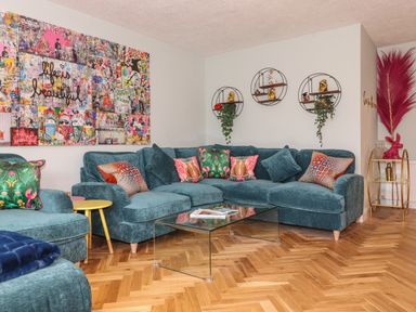 A living room with a teal sectional sofa with colorful cushions a glass coffee table wooden floor and wall art at Tipsy Bee in Bowness-On-Windermere