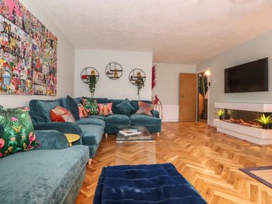 A living room with a blue sectional sofa colorful pillows a glass coffee table wall art and a wall-mounted television with an electric fireplace at Tipsy Bee in Bowness-On-Windermere