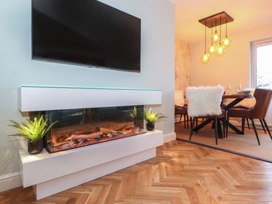 A modern living space featuring a wall-mounted television above a white electric fireplace with two potted plants on either side and a dining area with a table, four chairs with white fur covers, pendant lights and a window at Tipsy Bee in Bowness-On-Windermere