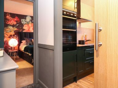 View of a kitchen with black cabinets and an oven next to a bedroom with a bed and floral wallpaper at Tipsy Bee Bowness-On-Windermere