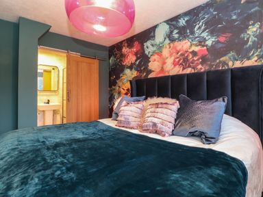 A bedroom with a large bed with multiple pillows a floral feature wall a wooden sliding door and a visible bathroom at Tipsy Bee in Bowness-On-Windermere