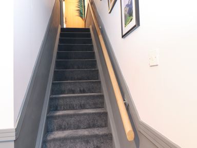 A staircase with dark carpet and wooden handrail with framed pictures on white walls at Tipsy Bee in Bowness-On-Windermere