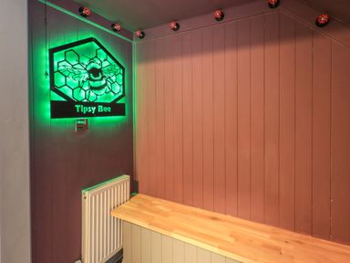 A corner with brown wooden paneling a radiator a wooden countertop and a green illuminated Tipsy Bee sign at Tipsy Bee in Bowness-On-Windermere