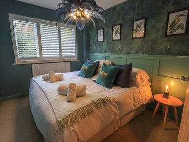 A bedroom with a double bed and rolled towels on it next to a small round table with a lamp and bird pictures on the wall at Tipsy Bee in Bowness-On-Windermere