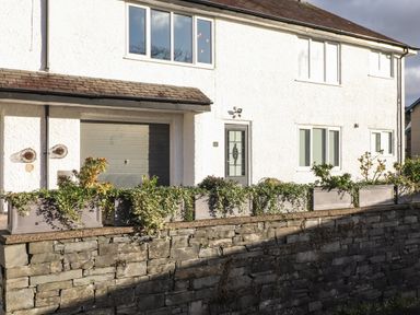 The front of a white house with a garage door a main door windows and planter boxes on a stone wall at Tipsy Bee in Bowness-On-Windermere