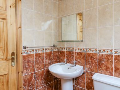 A bathroom with a white pedestal sink, mirror above it, tiled walls, a wooden door, and part of a toilet at 14 Sneem Leisure Village in Sneem County Kerry