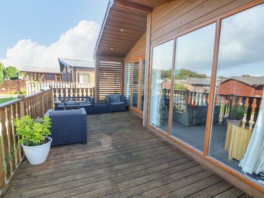 A wooden deck with outdoor seating including chairs and a glass table adjacent to a cabin with large windows at Gressingham 22 in South Lakeland Leisure Village