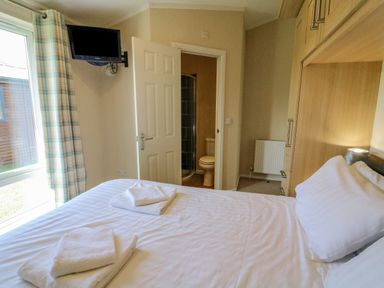 A bedroom with a double bed, folded towels on the bed, a window with plaid curtains, a wall-mounted TV, and a door leading to a bathroom at Gressingham 22 South Lakeland Leisure Village