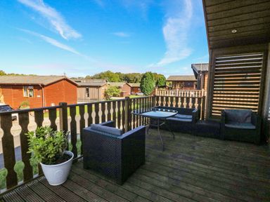 An outdoor wooden deck with dark rattan chairs a small table and a potted plant with wooden cabins and trees in the background at Gressingham 22 in South Lakeland Leisure Village