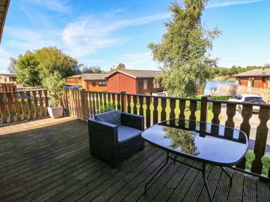 A wooden deck with a chair and glass table overlooking cabins and a lake at Gressingham 22 in South Lakeland Leisure Village