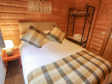 A bedroom with a double bed with plaid pillows and blanket wooden walls a lamp on a bedside table and a clothing rack at Leafy Hollow Lodge in Louth