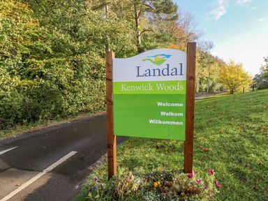 A wooden sign for Landal Kenwick Woods near a road surrounded by trees and grass