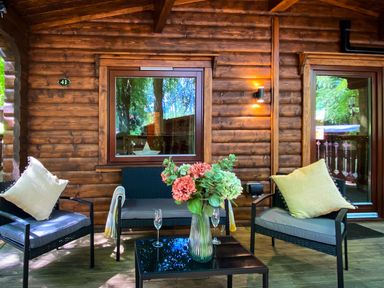 An outdoor wooden porch with chairs, cushions, a table with flowers, and windows at Leafy Hollow Lodge in Louth