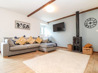 A living room with a grey sectional sofa and multiple pillows a wall-mounted TV a wood-burning stove with wood in a crate and a basket a light-colored rug on wooden floor at Pendeen in Mawnan Smith Near Falmouth