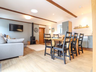 A combined living and dining area with a wooden dining table and black chairs a gray sofa mounted TV wood stove and kitchen cabinets at Pendeen in Mawnan Smith Near Falmouth