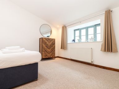 A bedroom with a bed, chest of drawers with a round mirror, window with curtains and decorative items at Pendeen Mawnan Smith Near Falmouth