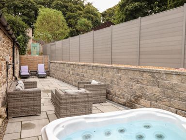 An outdoor patio area with wicker seating and a hot tub surrounded by a stone wall and fencing at Pendeen in Mawnan Smith Near Falmouth