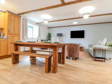 A dining area with a wooden table and benches beside a living room with a sofa and a wall-mounted TV at Budock in Mawnan Smith Near Falmouth