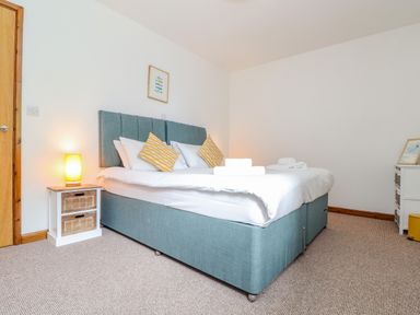 A bedroom with a blue upholstered bed white bedding yellow patterned pillows a small bedside table with a lamp and a framed picture on the wall at Budock in Mawnan Smith Near Falmouth