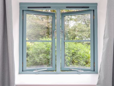 A blue framed open window with greenery outside and grey curtains on both sides at Budock in Mawnan Smith Near Falmouth
