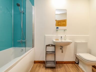 A bathroom with a bathtub shower combination a toilet a wall mounted sink a mirror and a small wooden shelf unit at Budock in Mawnan Smith Near Falmouth
