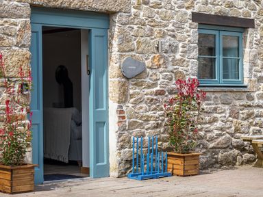 A stone building with a blue door and window two wooden planters with red plants and a blue object outside at Kerensa in Mawnan Smith Near Falmouth
