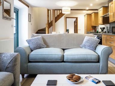 A living room with a blue sofa and pillows with a kitchen and wooden stairs in the background at Kerensa in Mawnan Smith Near Falmouth