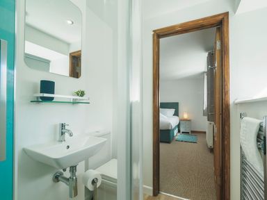 A small bathroom with a sink toilet and mirror next to a doorway leading to a bedroom with a bed nightstand and lamp at Kerensa in Mawnan Smith Near Falmouth
