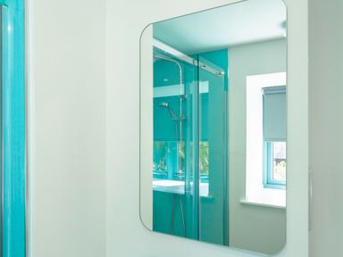 A bathroom mirror reflecting a shower area with turquoise wall panels and a window with a roller blind at Kerensa in Mawnan Smith Near Falmouth