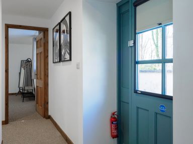 A hallway with a blue door and fire extinguisher next to a doorway leading to a room with a standing mirror and clothes rack at Kerensa in Mawnan Smith Near Falmouth
