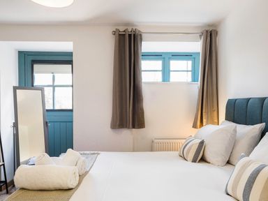 A bed with white linens and striped pillows near windows with curtains and a standing mirror in a bedroom at Kerensa in Mawnan Smith Near Falmouth