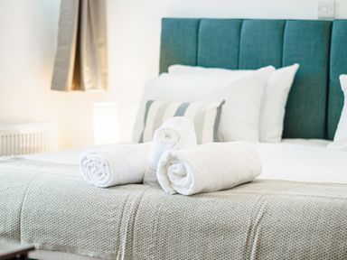 A bed with a blue headboard and white pillows with rolled white towels on a beige blanket at Kerensa in Mawnan Smith Near Falmouth