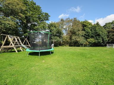 A grassy backyard with a trampoline a wooden playset with slide and two small soccer goals surrounded by trees at Newlyn in Mawnan Smith Near Falmouth