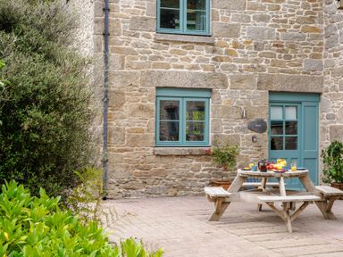 An outdoor patio with a wooden picnic table set for breakfast next to a stone building with blue windows and door at Newlyn in Mawnan Smith Near Falmouth